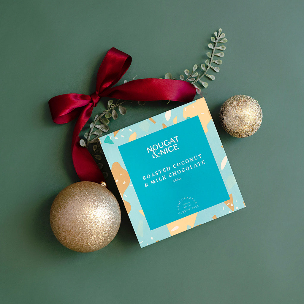 A 200g box of Roasted Coconut & Milk Chocolate Nougat, adorned with a red ribbon and decorative elements, against a green background.
