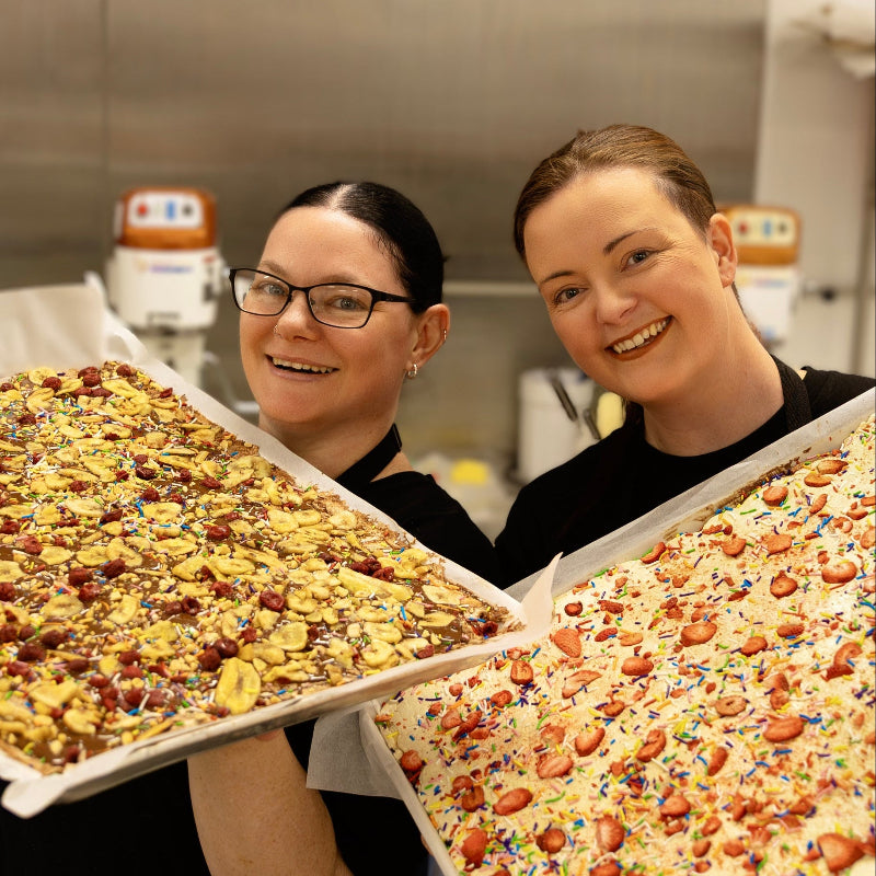 Two people holding large trays of food in a kitchen setting